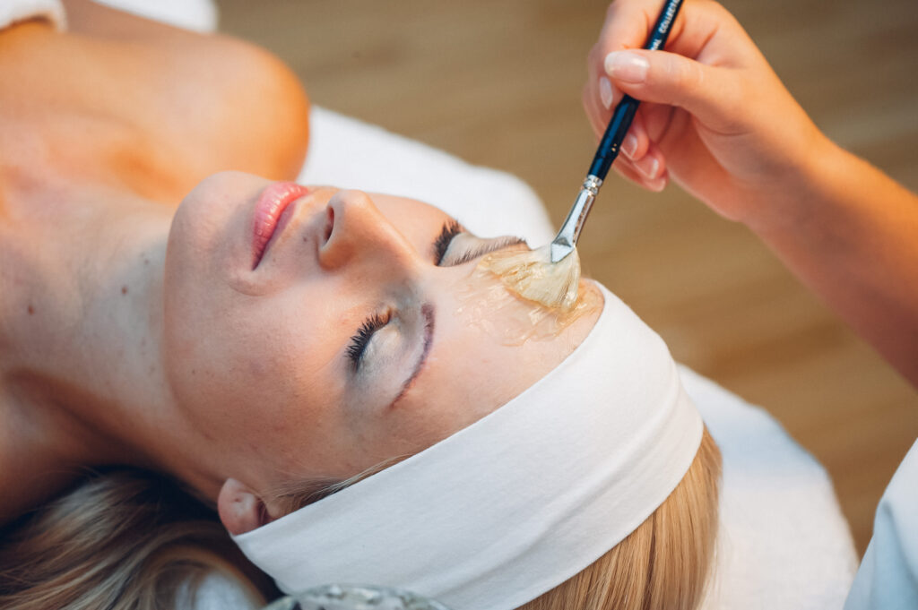 Eine Frau mit geschlossen Augen erhält eine Gesichtsmaske, die mit einem Pinsel aufgetragen wird.