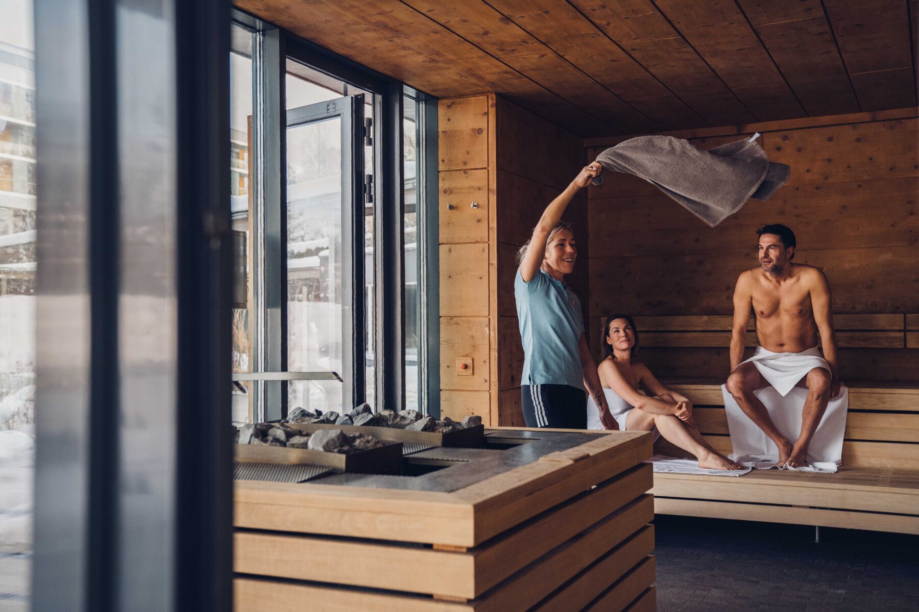Zwei Saunagäste und eine Saunawartin, die gerade mit dem Handtuch die Hitze verteilt in der Saunakabine. Hin Hintergrund das eingeschneite Hotel im Winter.