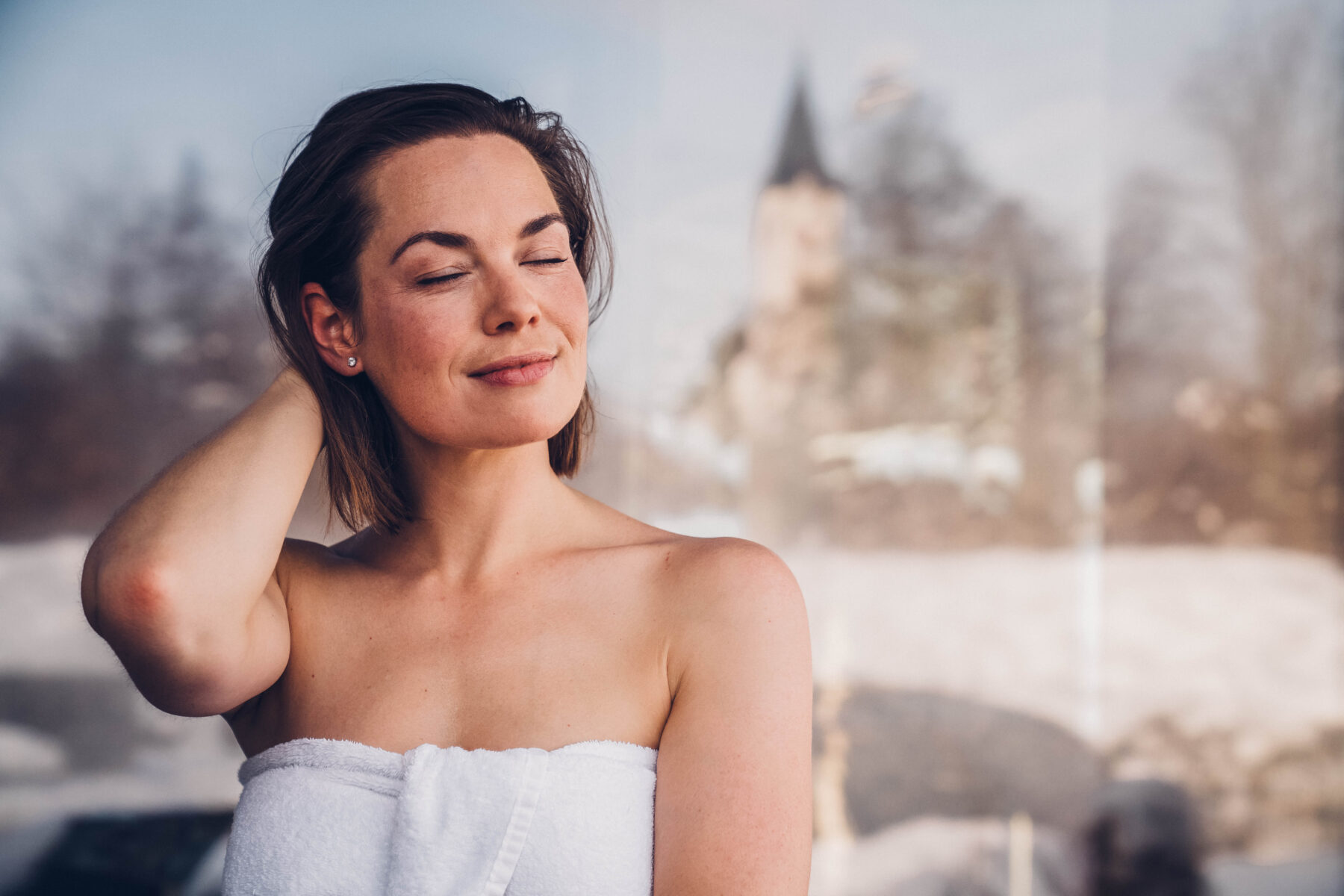 Eine Dame mit einem Handtuch bekleidet, welche sich mit einer Hand in die Haare fährt. mit einem entspannten und glücklichen Blick. Im Hintergrund das verschneite Bad Zell zu erkennen.