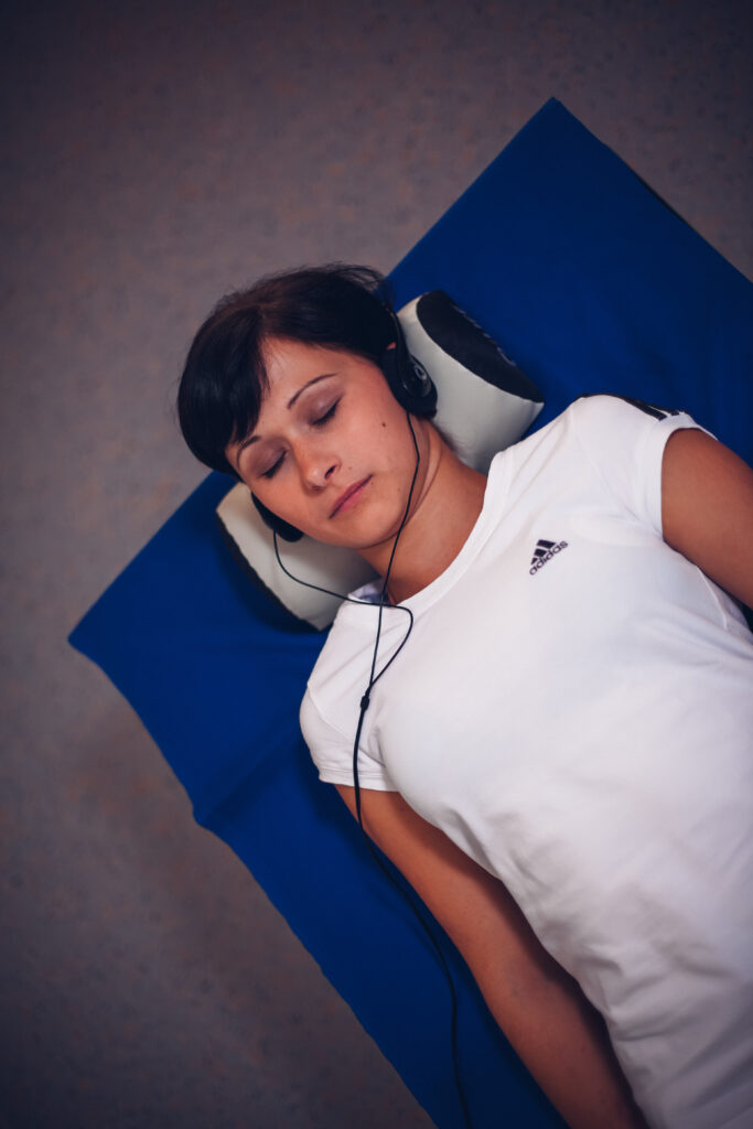 Eine Frau die auf einer iLife-SOMM-Liege liegt mit Polster und Kopfhörer auf den Kopf hat.