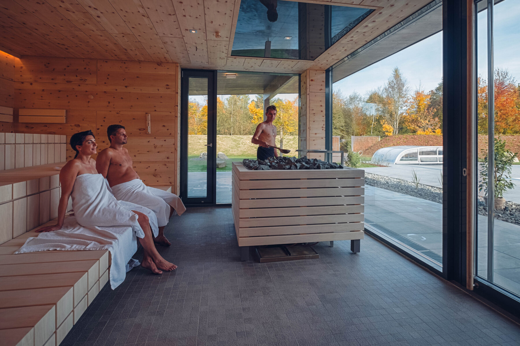 Sauna mit einem Saunameister, der gerade einen Aufguss macht und zwei Personen auf er Bank sitzend am entspannen.