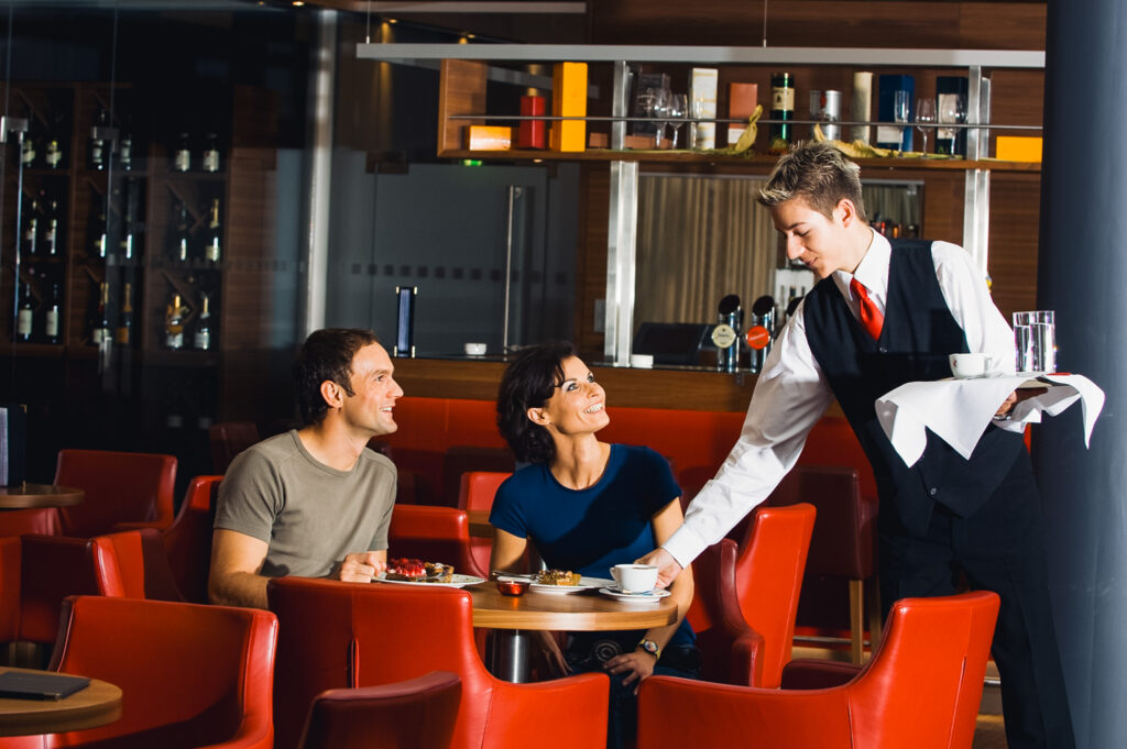 Man sieht einen Kellner, der gerade dabei ist, an einem Tisch mit 2 Personen den Kaffee einzustellen. Im Hintergrund ist die Hotelbar zu erkennen.